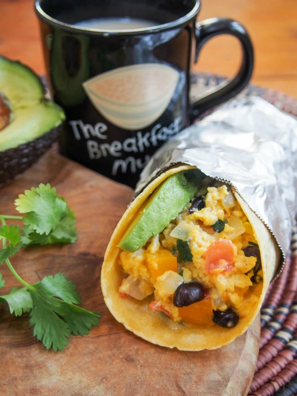 breakfast burrito resting on wooden board with cilantro and avocado to side and mug behind