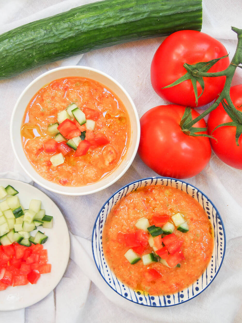 two bowls of gazpacho from overhead with plate with chopped cucumber and tomato to side