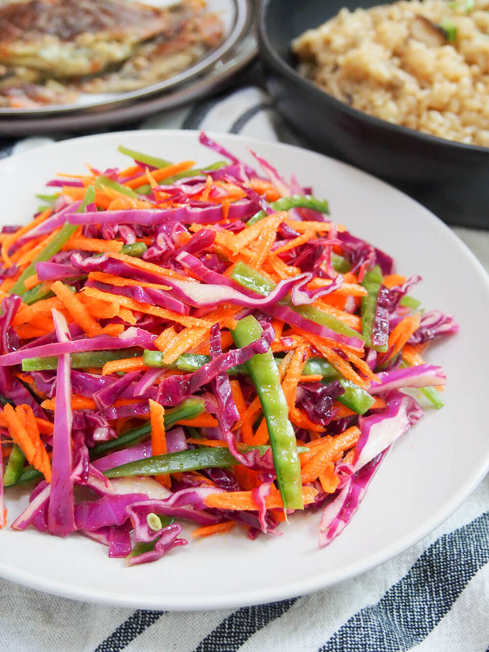 This rainbow slaw has only a few ingredients, but is both bright on your plate and in your mouth. Easy, crunchy and flavorful, it's versatile and great for potlucks and more. plate of rainbow slaw with other dishes behind
