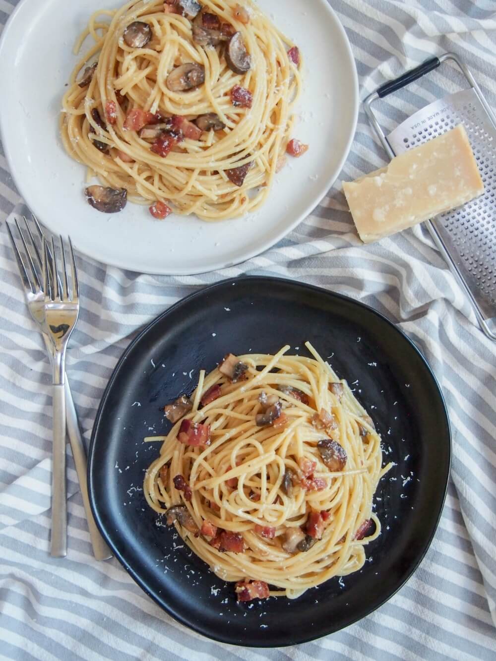 two plates of spaghetti alla carbonara with mushrooms from above, with forks to one side and grater and cheese to other
