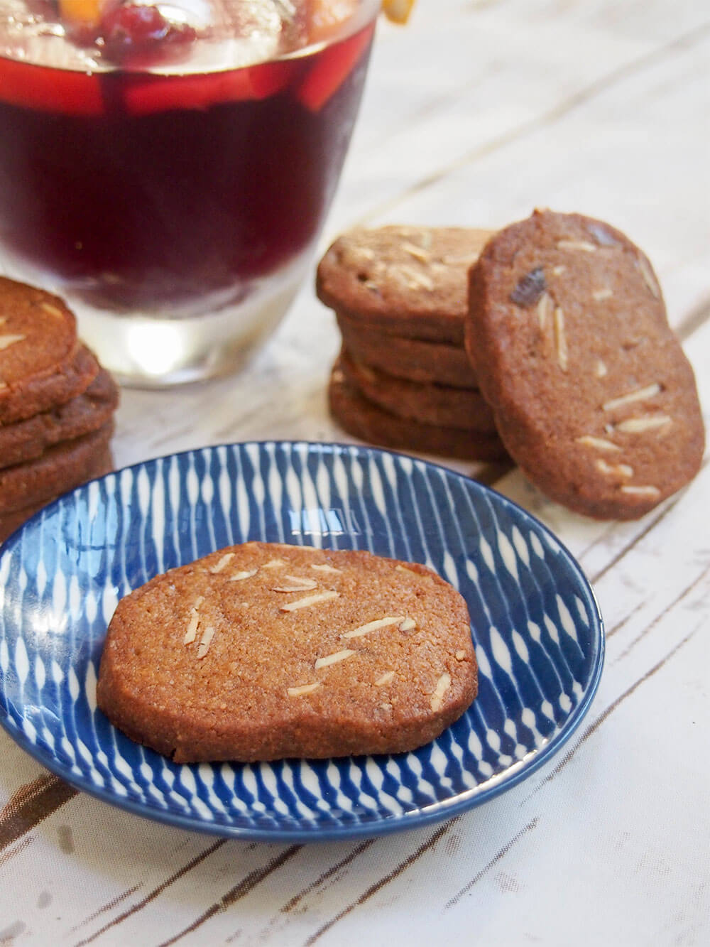 Brunkager - Danish Christmas cookies - with one cookie on a plate in front of a stack and glass on cranberry sangria behind