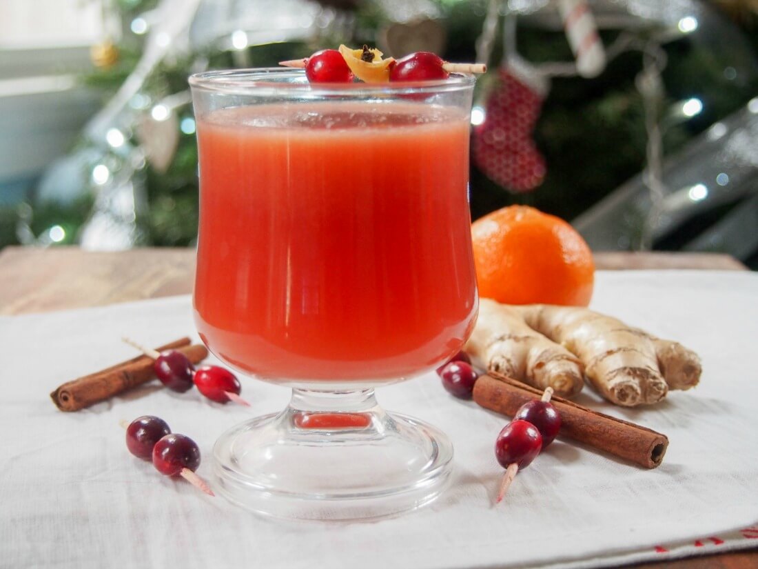 Warm spiced cranberry cocktail or mocktail with cranberries on cocktail sticks and spices around glass in front of Christmas tree
