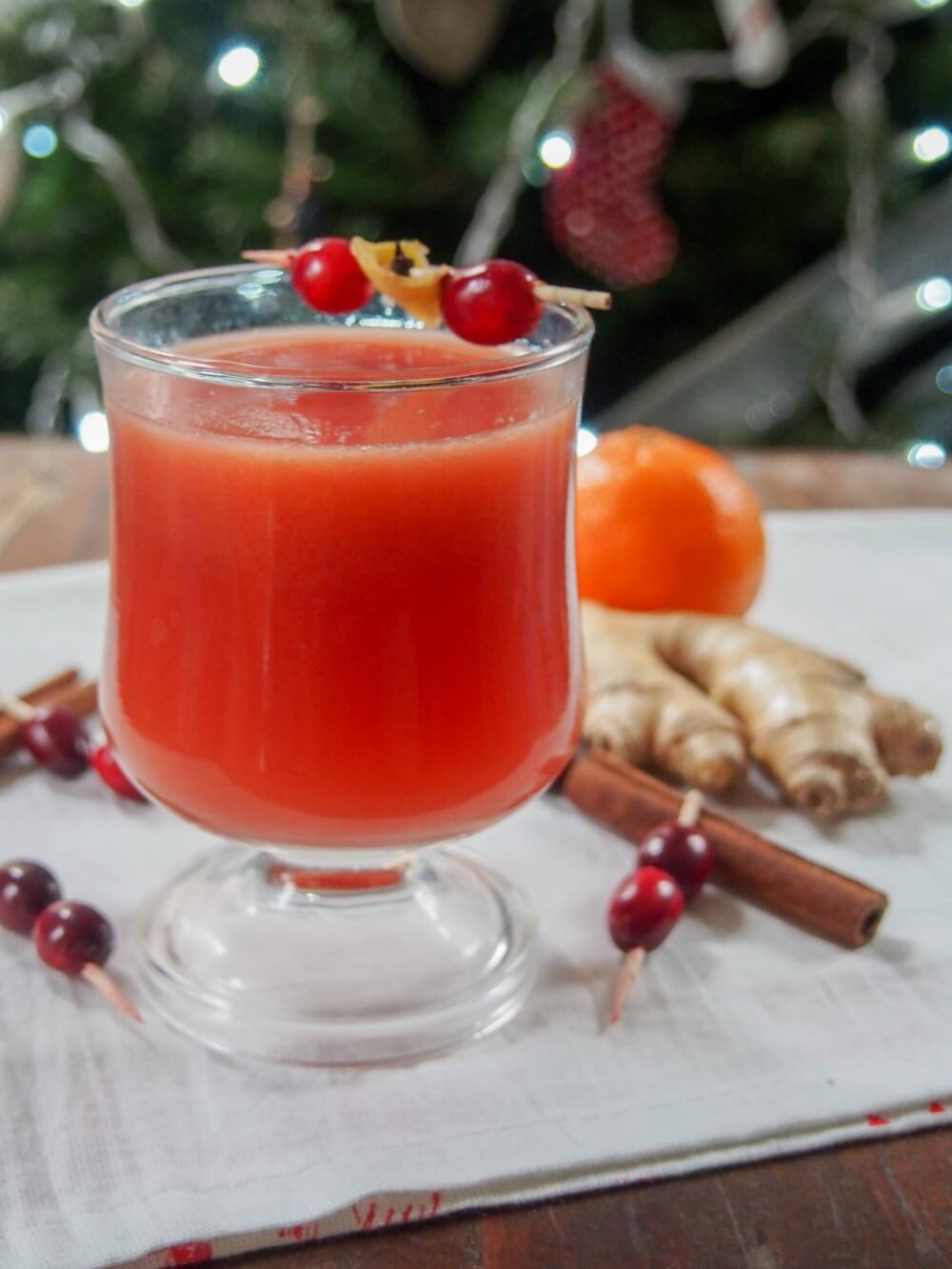 glass of warm spiced cranberry cocktail in front of Christmas tree with spices around glass