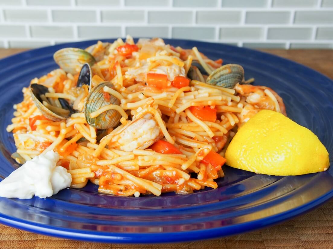 Fideua is a Catalan fish and seafood paella made with short pasta and a tomato-pepper and stock sauce base. Deliciously comforting. serving of fideua on plate with lemon slice to one side and aioli on other