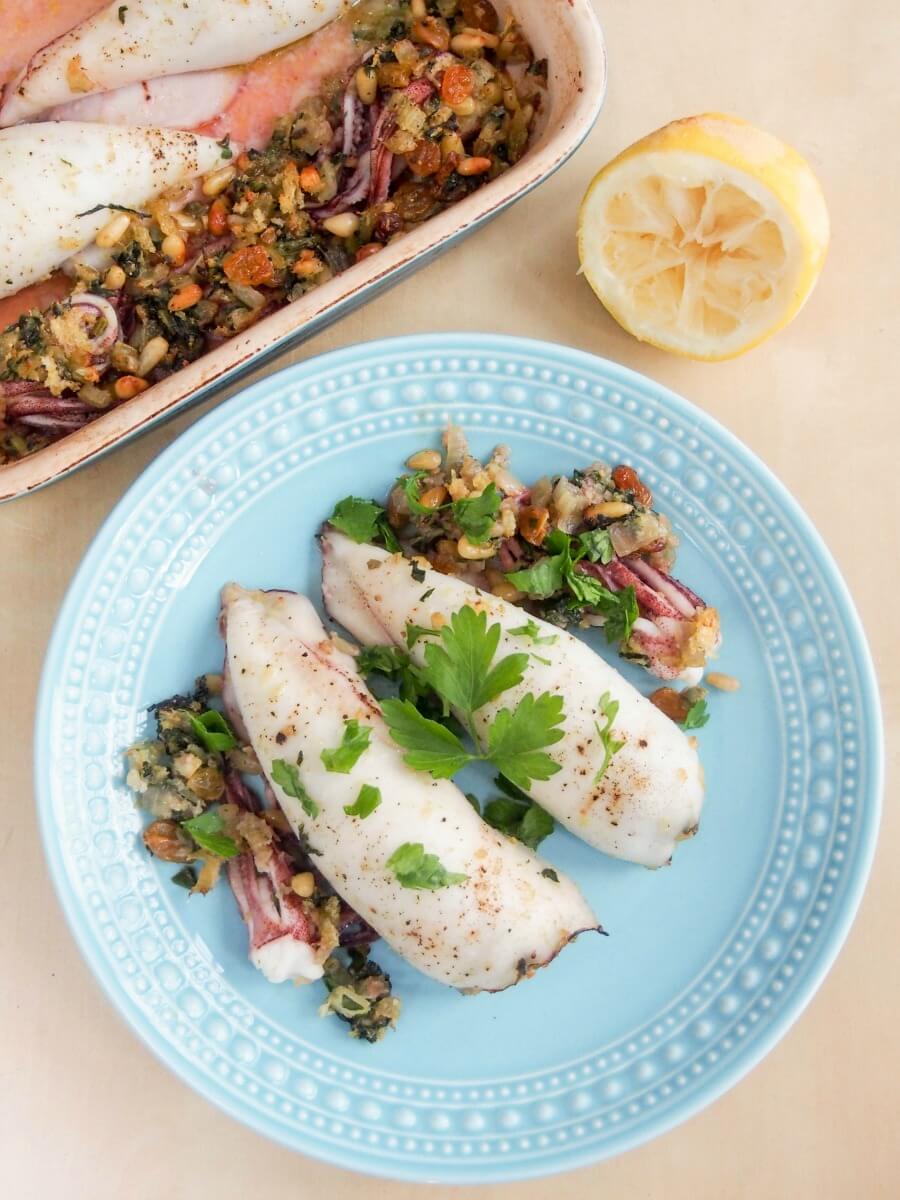 two pieces of Sicilian style stuffed squid on plate from overhead with cut lemon and additional squid in baking dish above