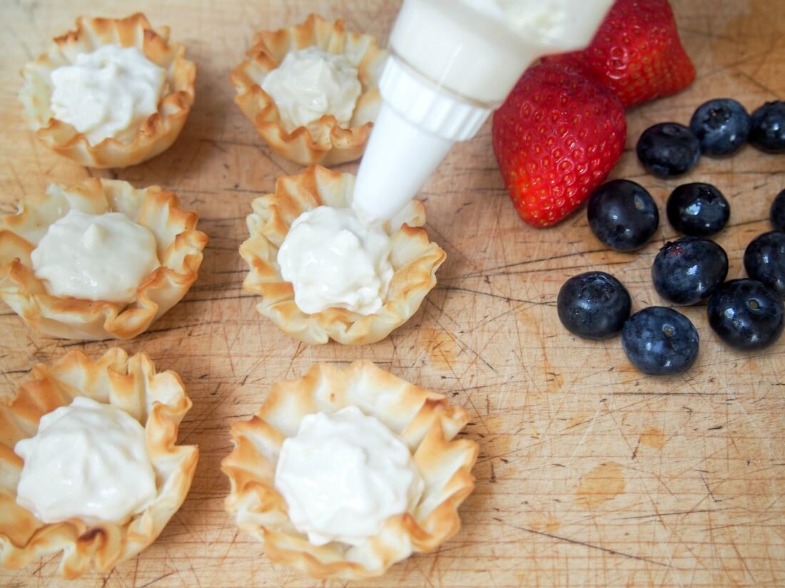 filling phyllo cups with lemon cheesecake cream with berries sitting to side