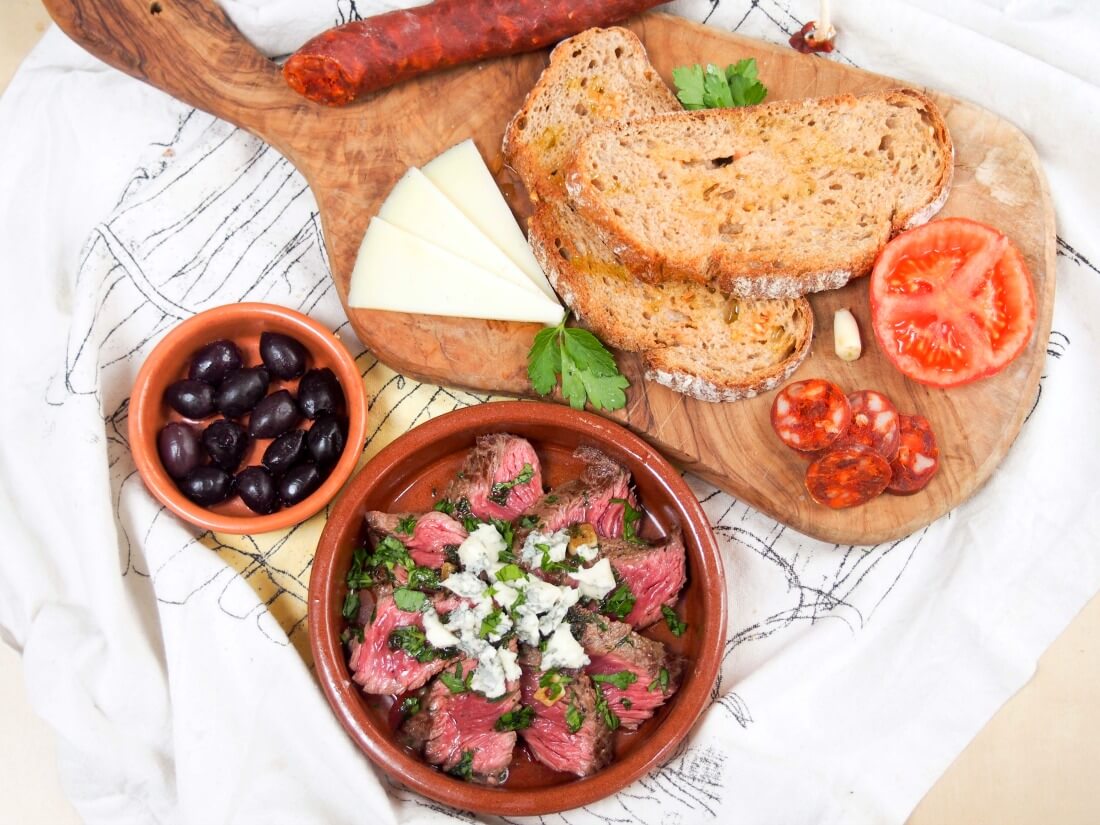 overhead shot of various tapas - tomato bread, cheese, chorizo, olive and steak dish
