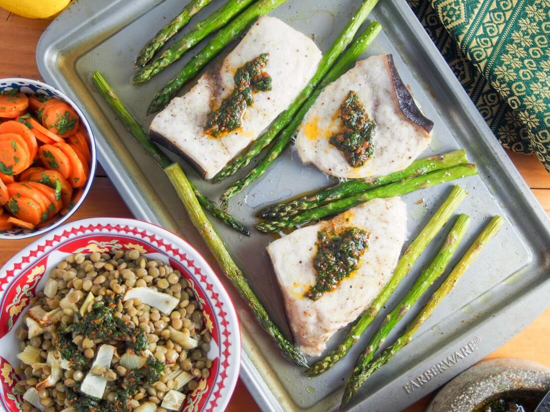 Baked swordfish and asparagus topped with chermoula and bowls of lentils and carrots to side, from overhead
