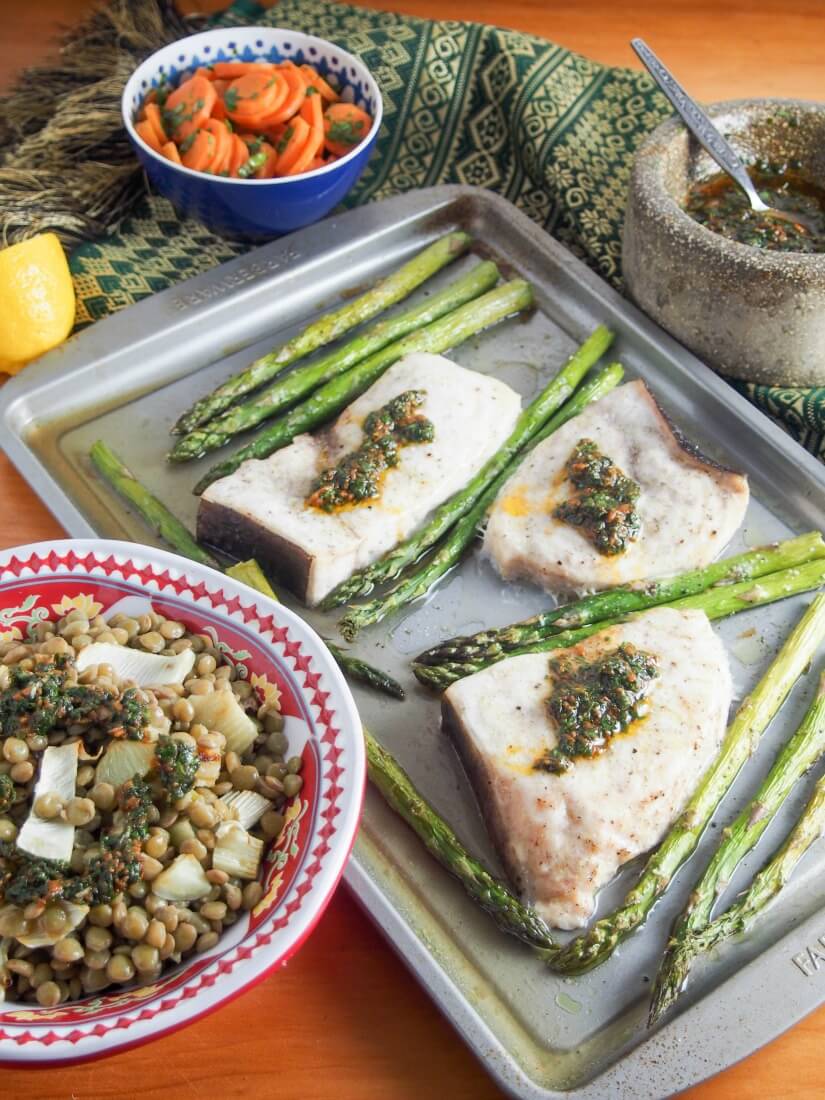 baked swordfish and asparagus on baking sheet with sides of lentils and carrots in bowls and extra sauce to side