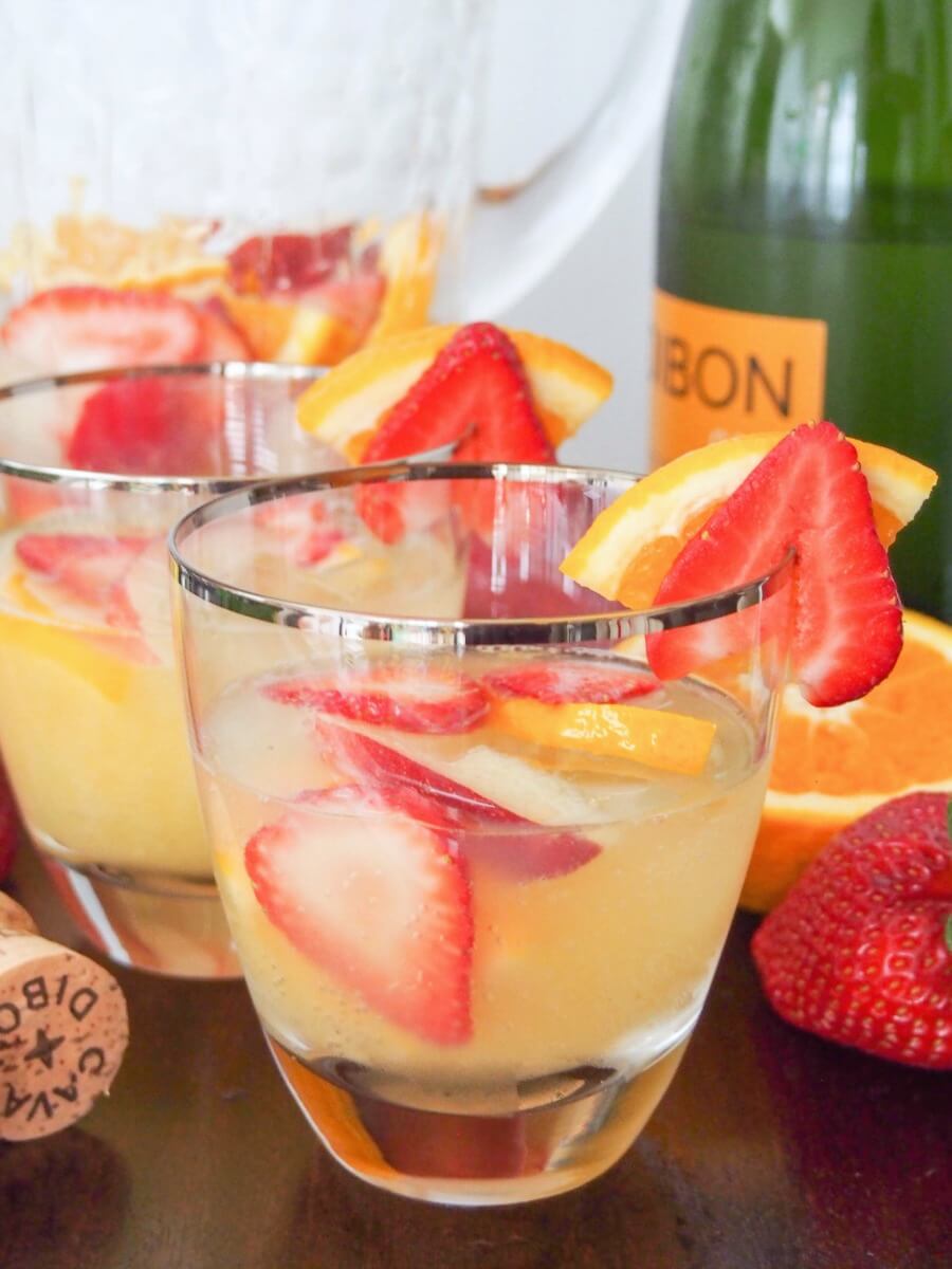 Sangria is a summer classic, but cava sangria is that bit more special. Light, refreshing and a wonderful mix of fruit and cava's bubbles. Quick to make, it's an easy cocktail perfect for any occasion from summer BBQs to holiday parties. cava sangria glass with pitcher behind and cava bottle