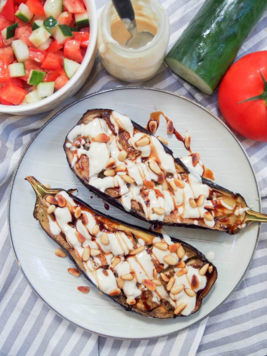 Roasted eggplant with tahini is a delicious appetizer or mezze dish that you'll find in Israel and other parts of the region. It's incredibly easy to make and full of fantastic flavor. Two halves of roasted eggplant with tahini on plate with Israeli salad to side