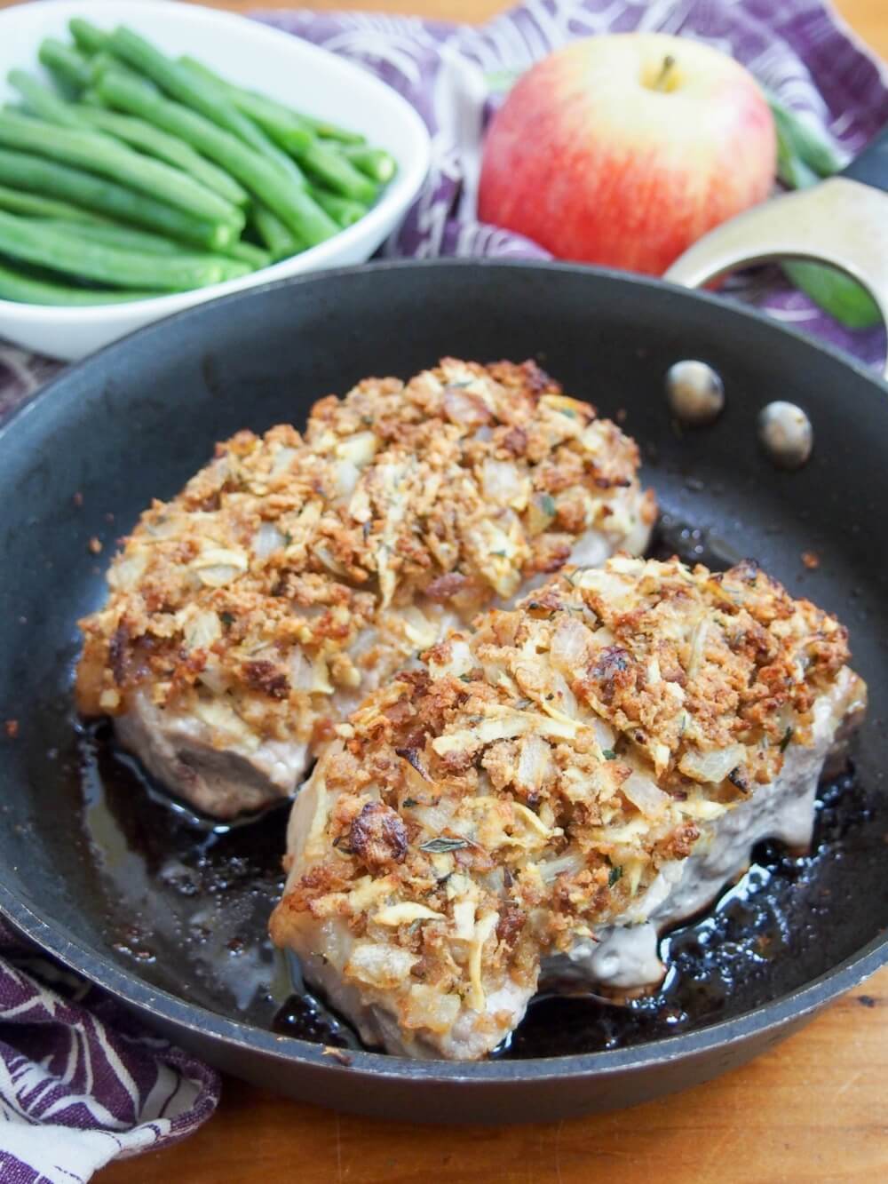 two apple stuffing crusted pork chops in skillet with bowl of green beans and an apple behind