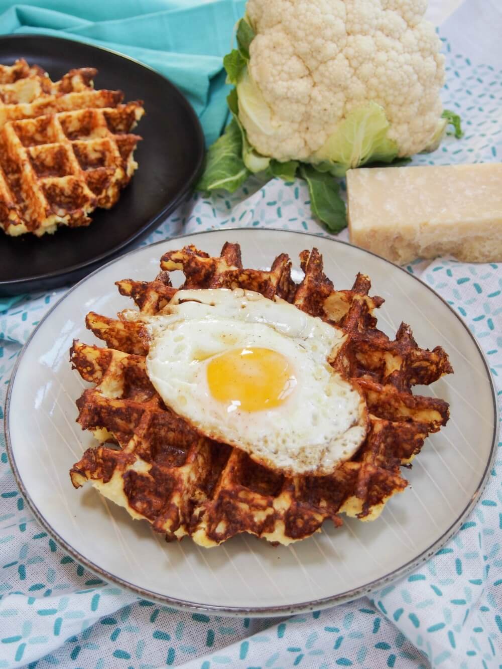These cauliflower waffles need only 5 ingredients and are quick to make. They make a tasty, low carb and gluten free any-time snack/ light meal. Cauliflower waffles with main waffle topped with egg, other not, and cauliflower and cheese behind plate