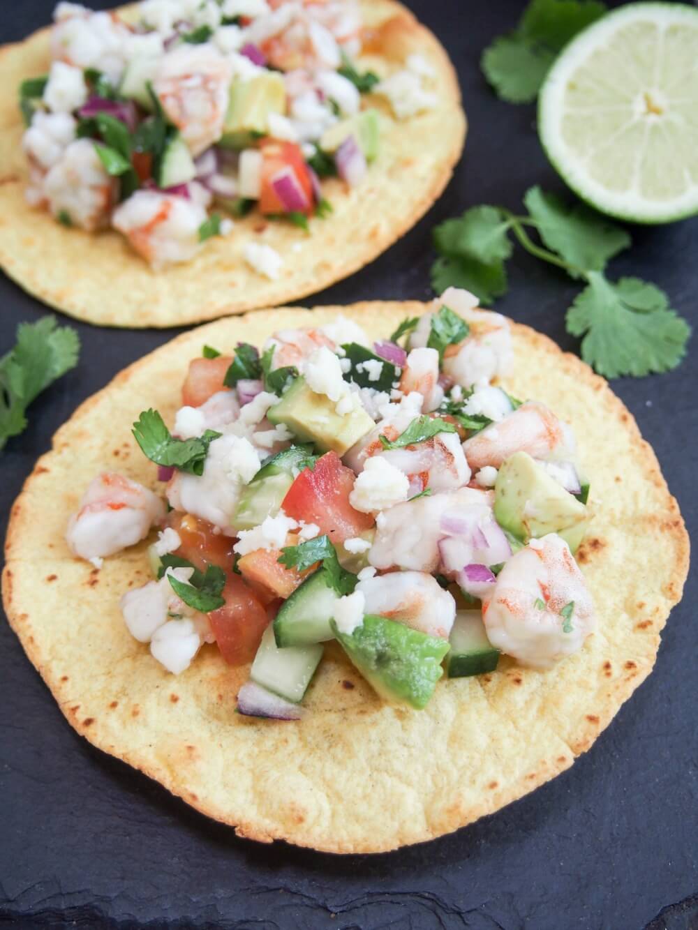Two shrimp ceviche tostadas sitting on slate board with lime and cilantro decoration to side