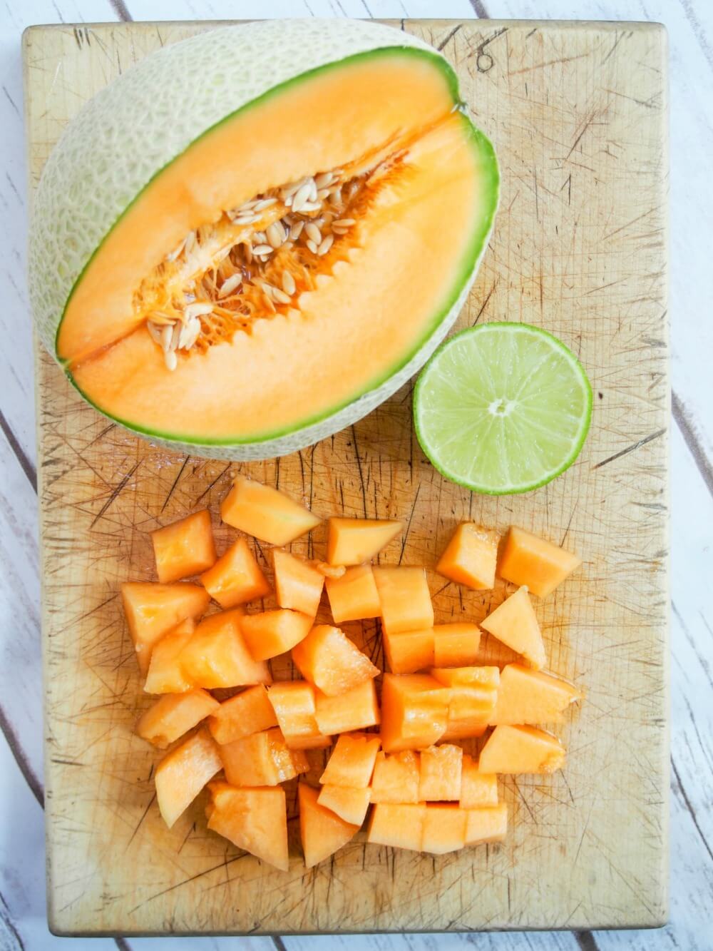 part of a cantaloupe diced up at bottom of cutting board with part lime and rest of cantaloupe above