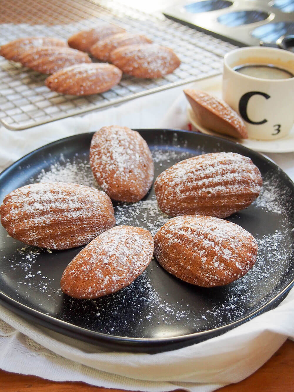 gingerbread madeleines on plate with more on cooling rack behind and one on saucer with coffee cup