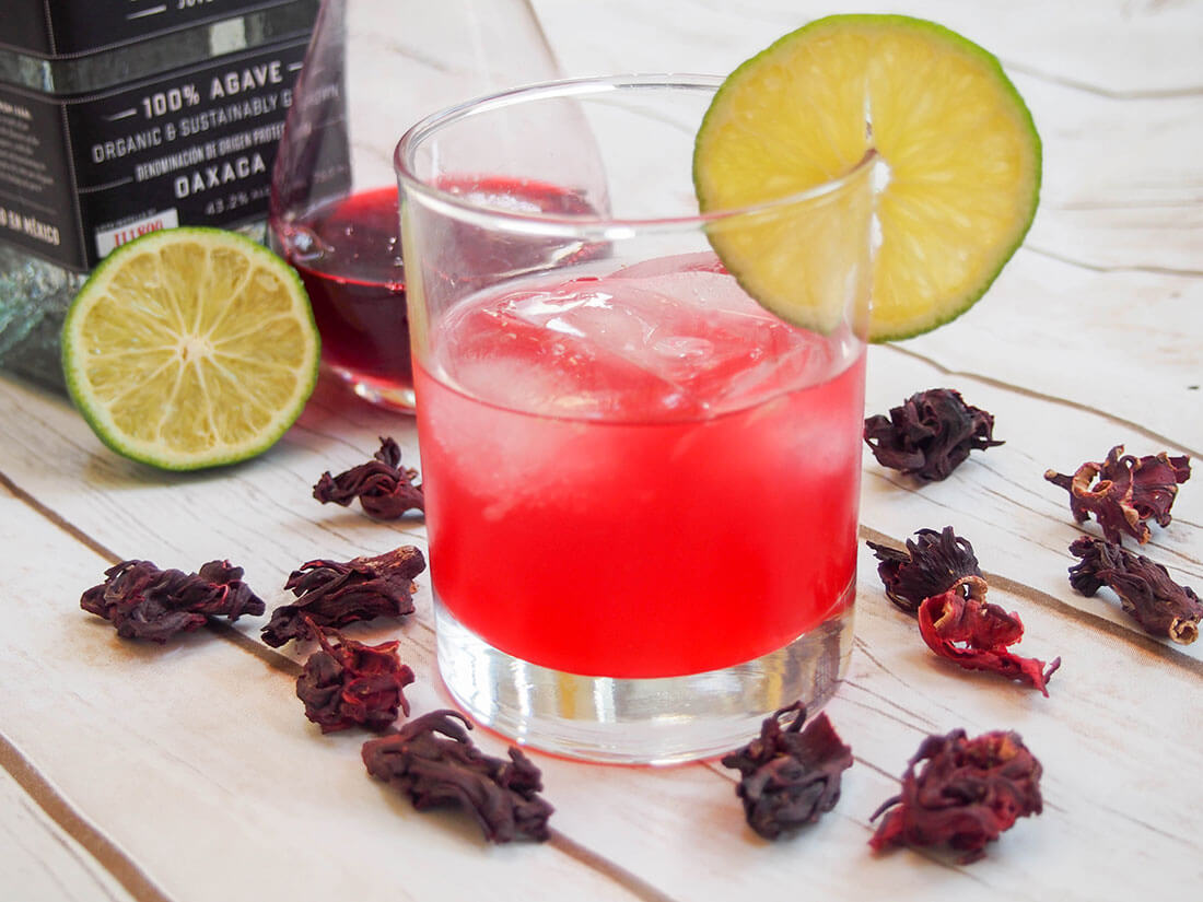 hibiscus mezcal sour with hibiscus flowers around glass and lime and bottles behind