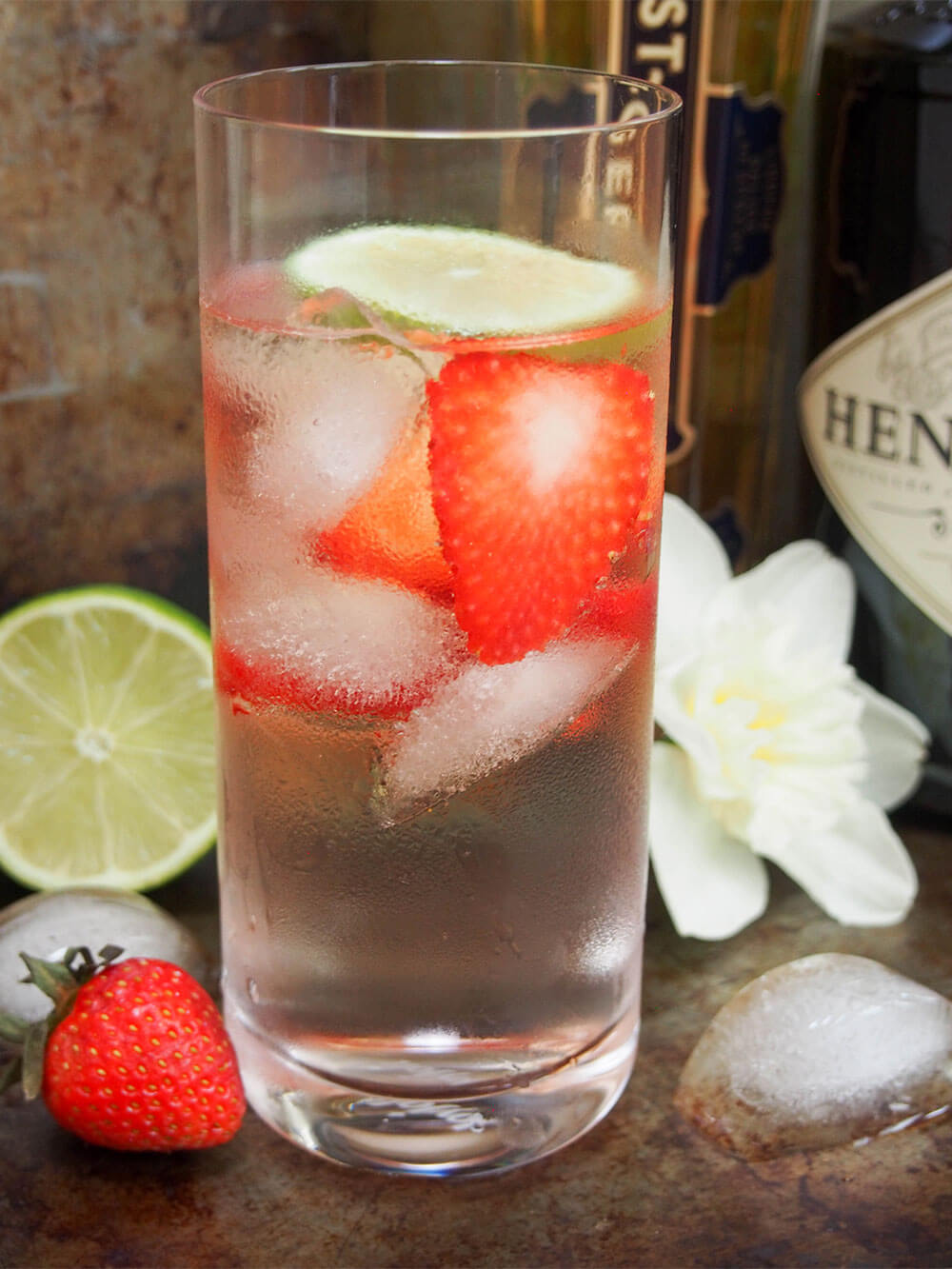 glass of finished cocktail with bottles behind and strawberry and cut lime to side