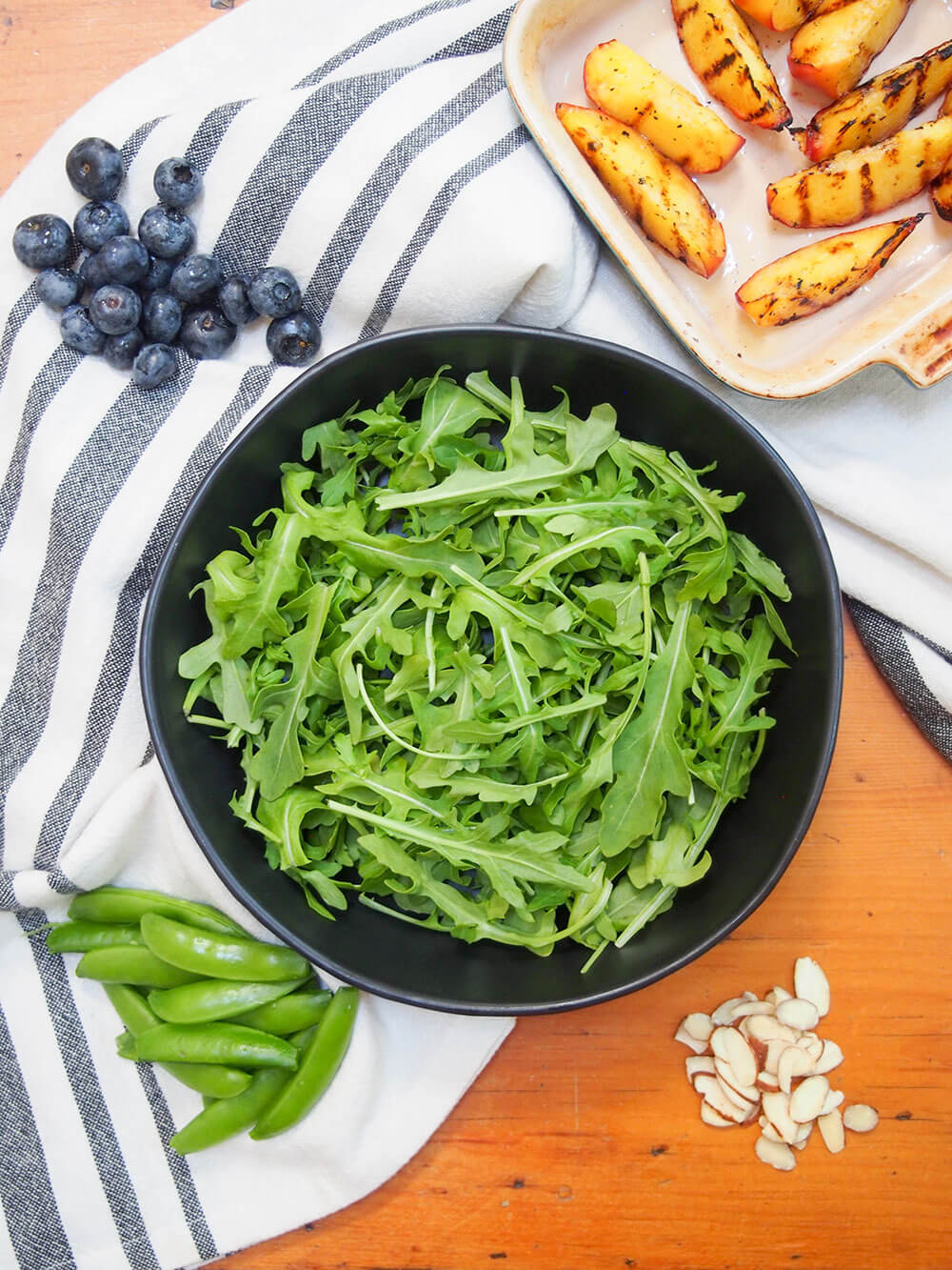 arugula in bowl with grilled peaches to one side, blueberries, snap peas and almonds in small piles around bowl