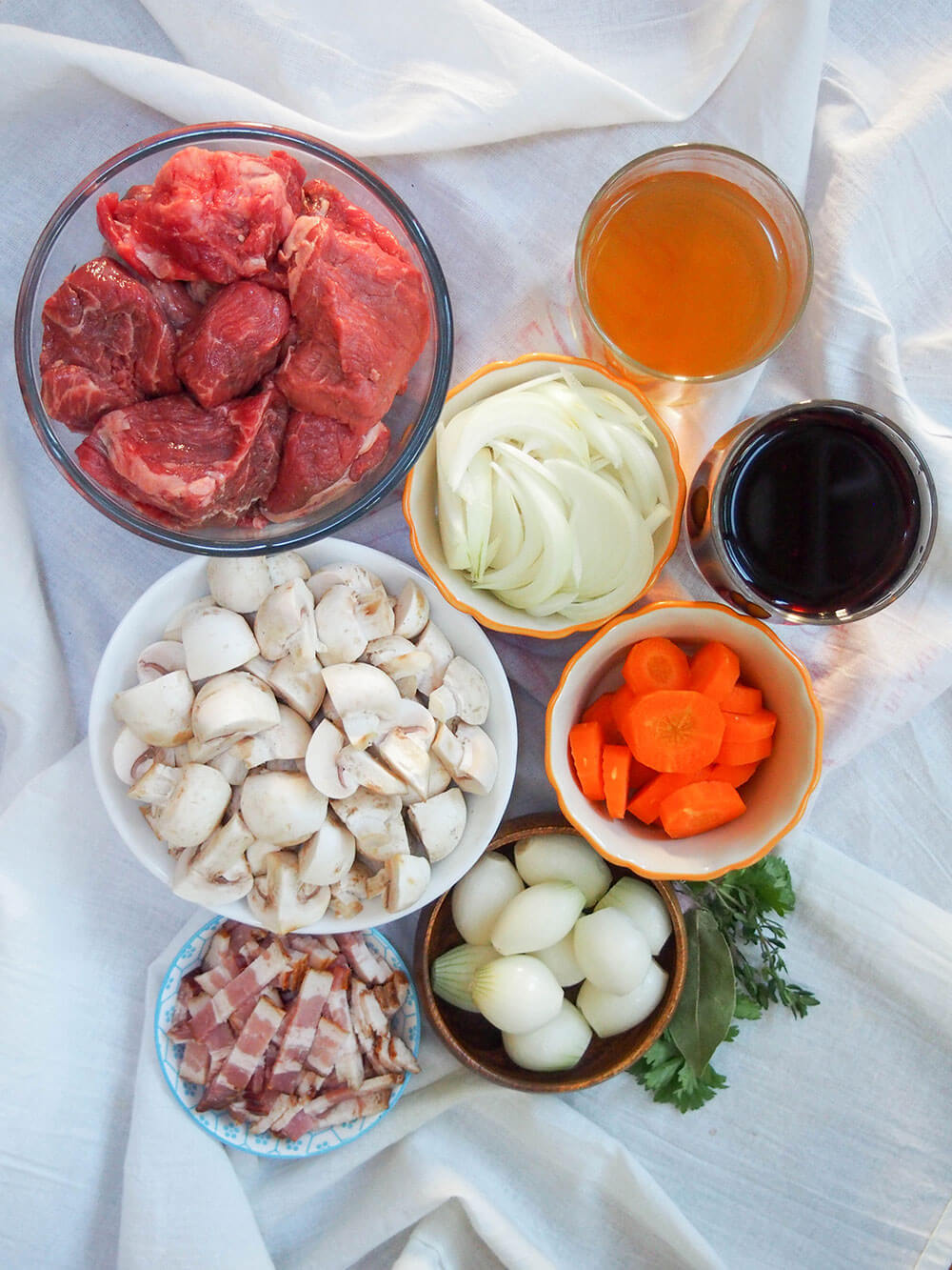Beef bourguignon ingredients laid out, with each in separate bowls or glasses and herbs to side