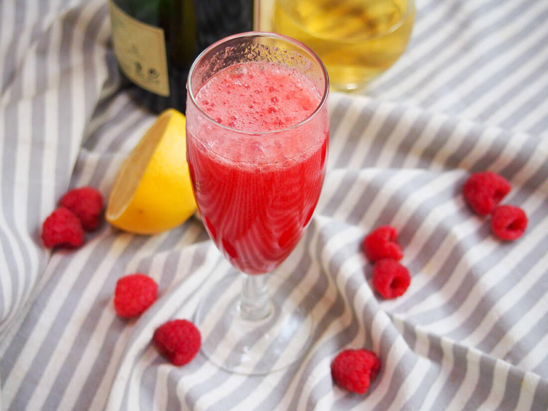 lemon raspberry bellini glass with bottles behind and lemon and raspberries around glass