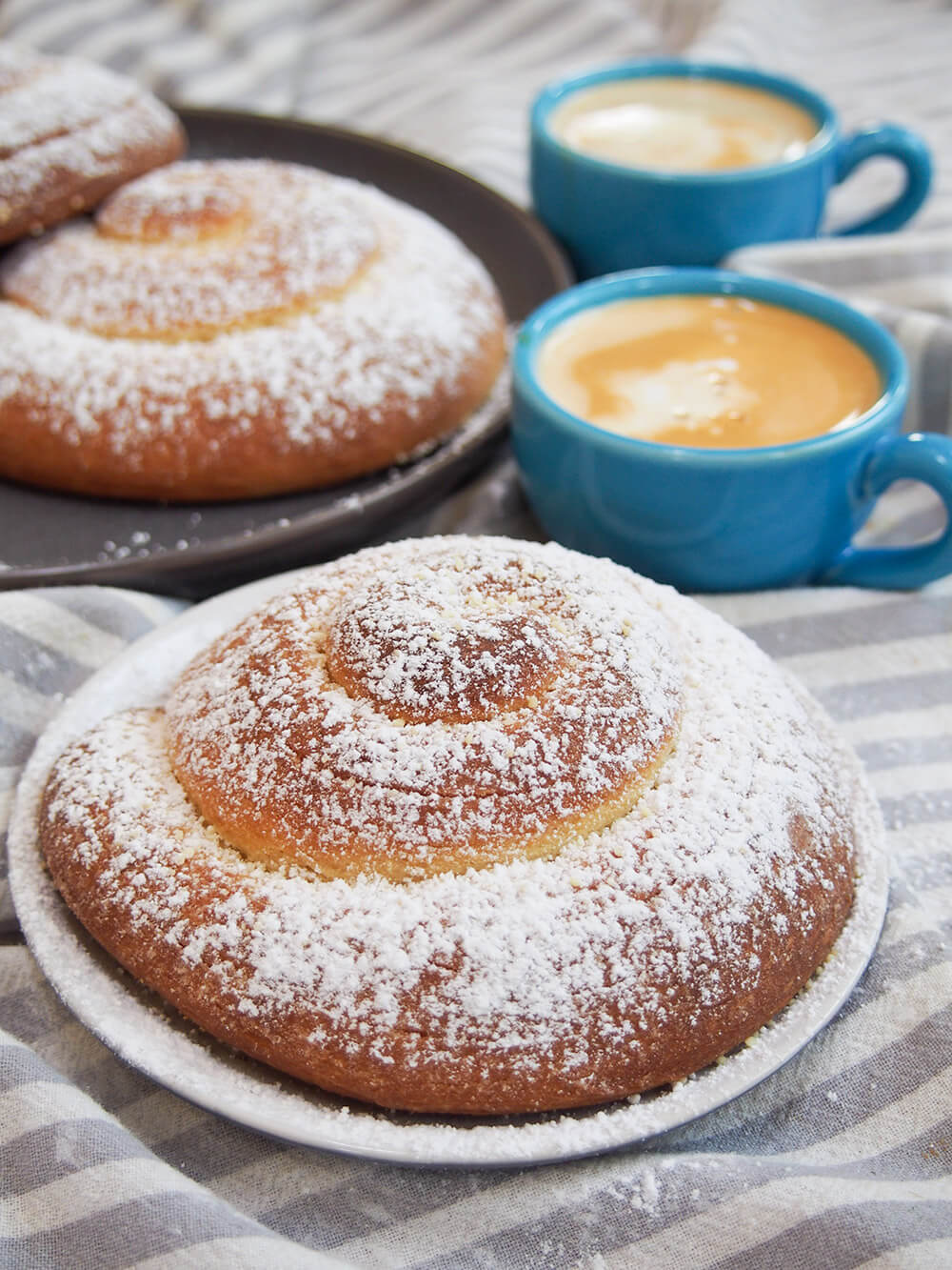 Pan de Mallorca (Puerto Rican sweet roll) on plate with additional rolls and cups of coffee behind