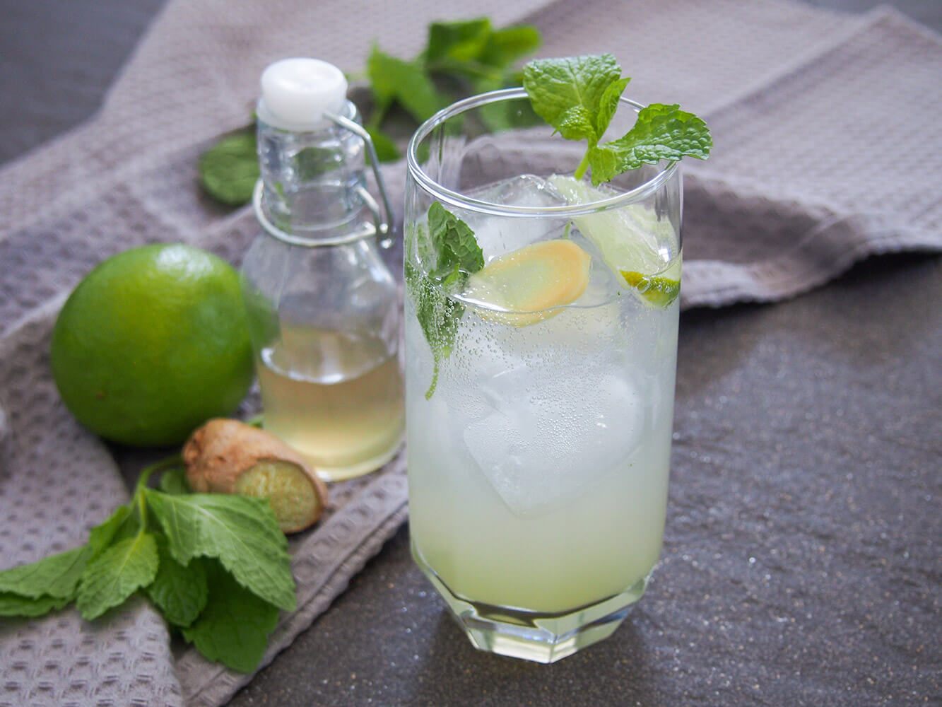 glass of ginger mojito with bottle of syrup, lime, mint and ginger to side