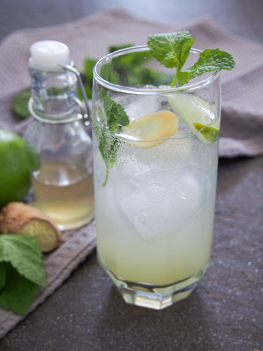 glass of ginger mojito with mint, lime and ginger garnish and bubbles showing and bottle of syrup behind