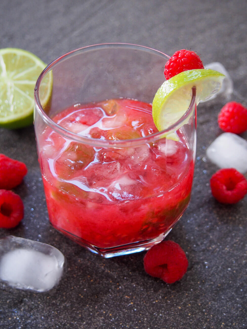 glass of raspberry caipirinha with ice, raspberries and lime around it
