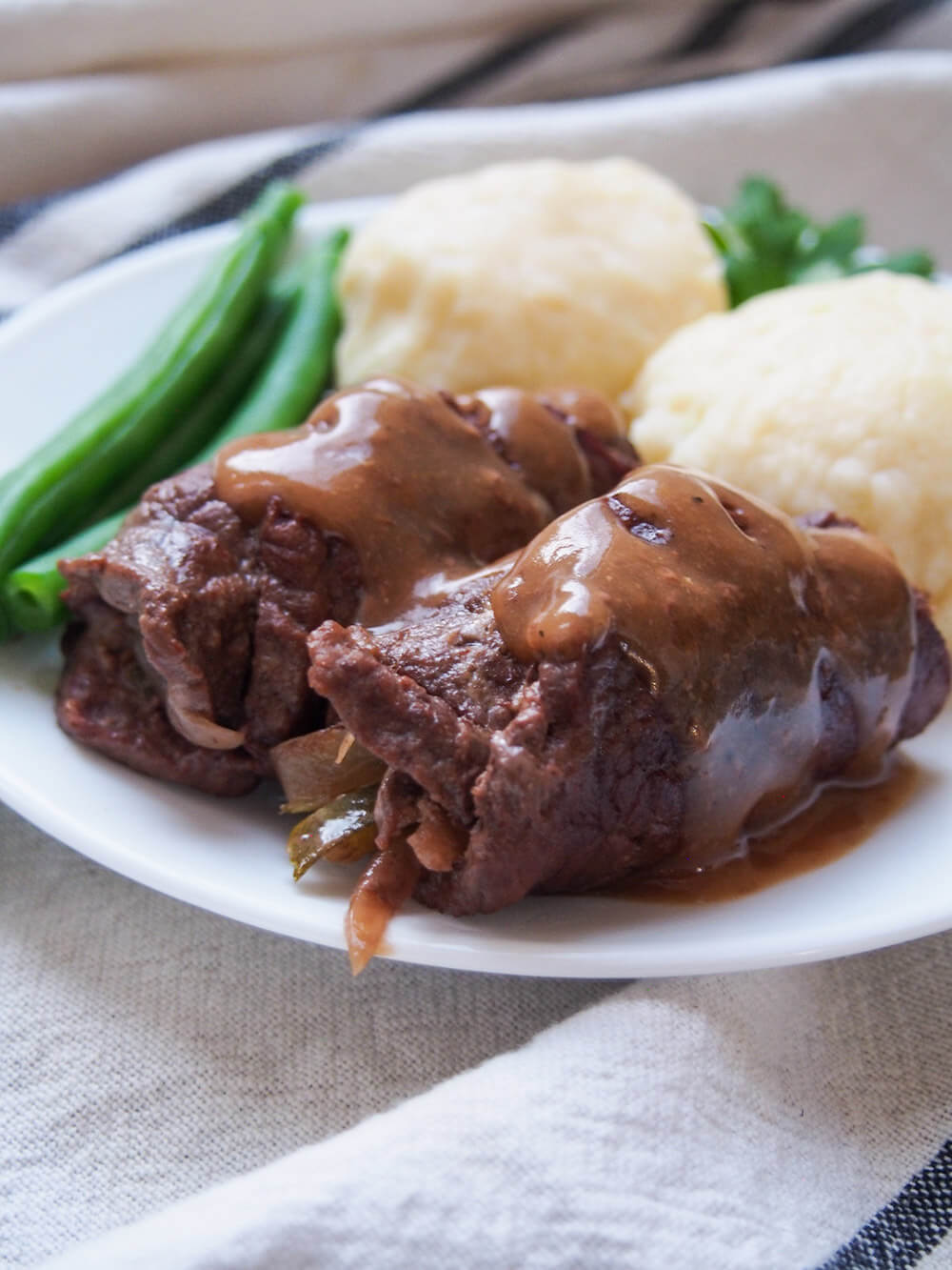 side view of beef rouladen topped with gravy with dumplings and green beans behind