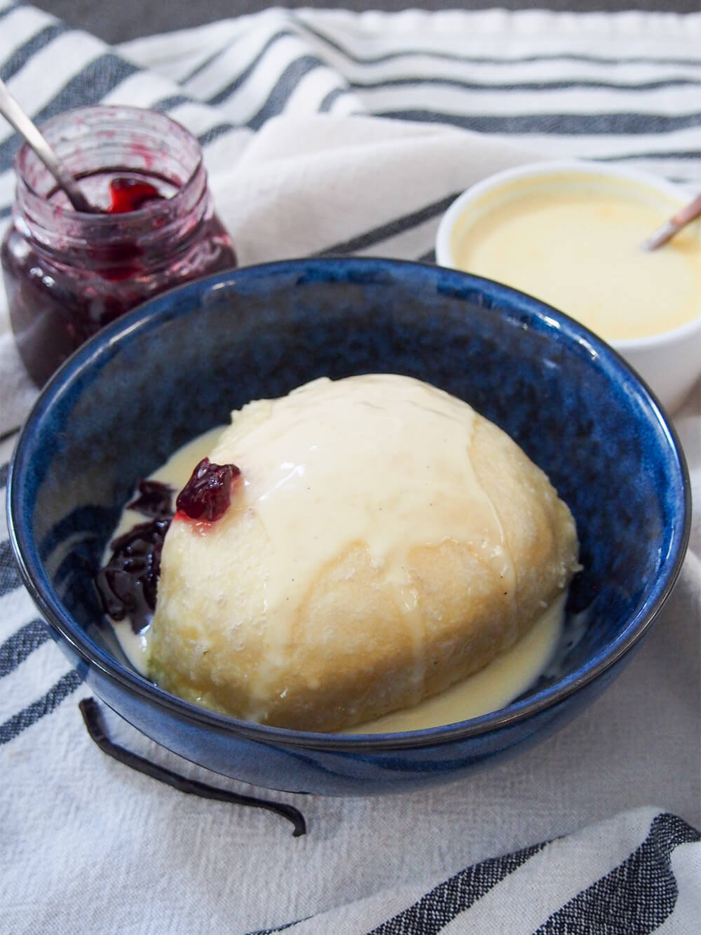 Dampfnudel is a steamed German sweet dumpling that's almost like a sweet bread, served with a luscious vanilla custard and often fruit compote. It's hearty but wonderfully comforting and delicious. dampfnudel served with vanilla sauce over top and jam to side with jam jar and bowl of sauce behind