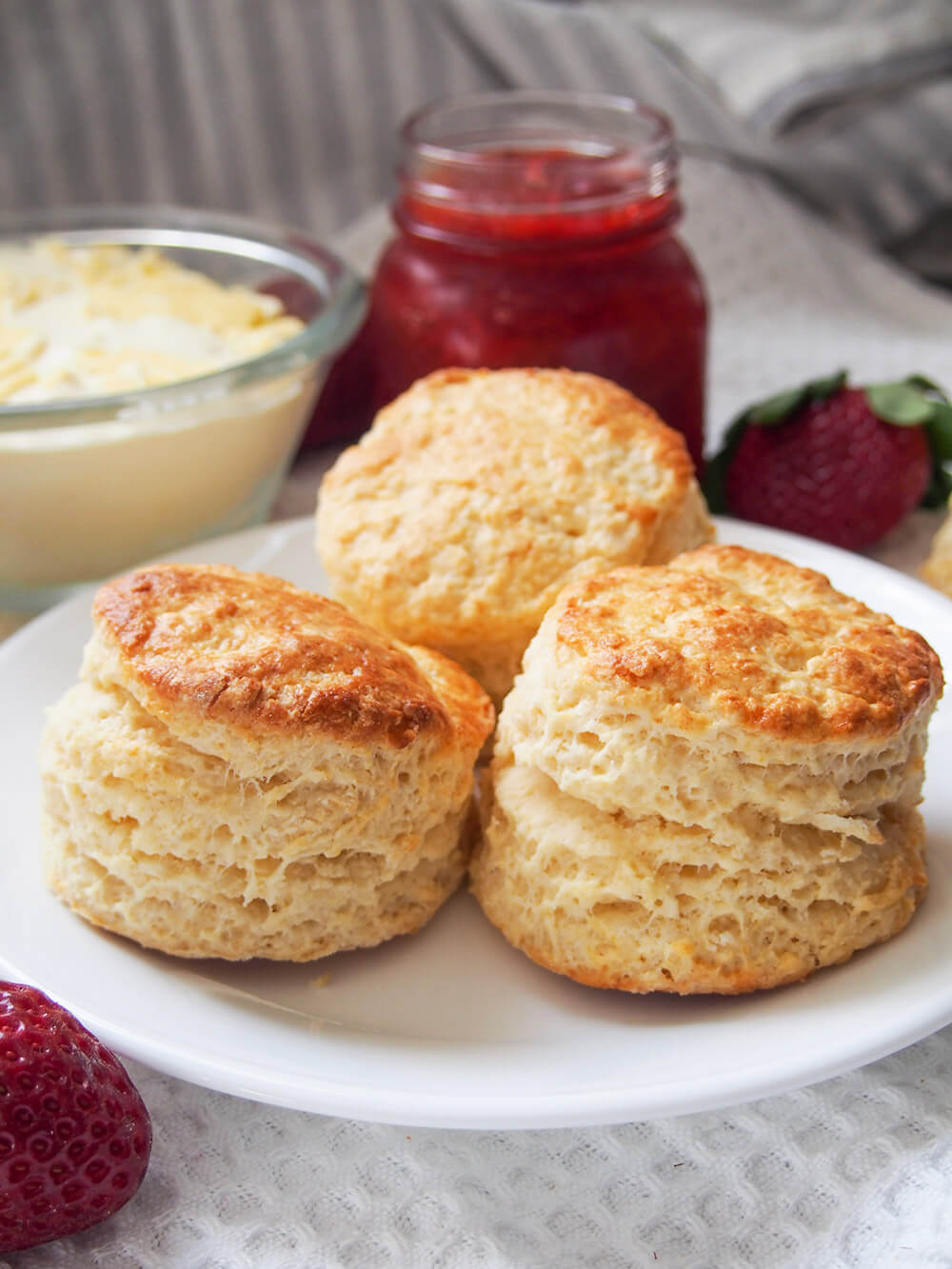 British scones are a classic treat to enjoy as part of a 'cream tea' with jam and cream. They are easy to make, wonderfully soft and gently sweet. Delicious in every bite. three British scones on plate with jam and cream behind