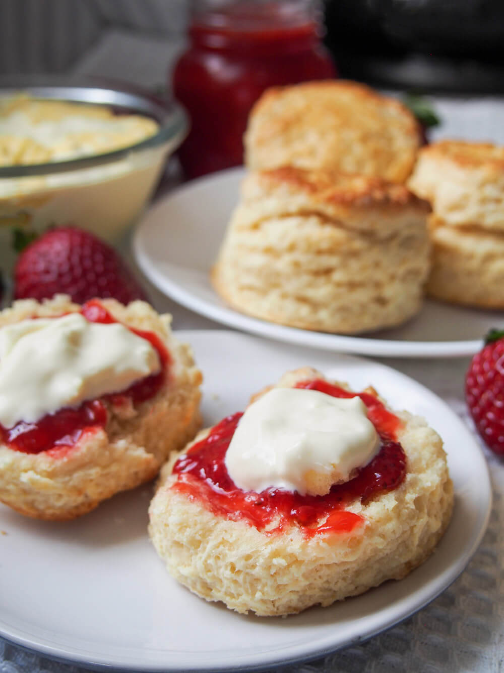 British scones are a classic treat to enjoy as part of a 'cream tea' with jam and cream. They are easy to make, wonderfully soft and gently sweet. Delicious in every bite. a scone with jam and cream with more scones, jam and cream dishes behind