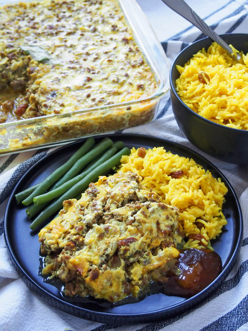 bobotie on plate with yellow rice and beans and rest of bobotie in dish behind