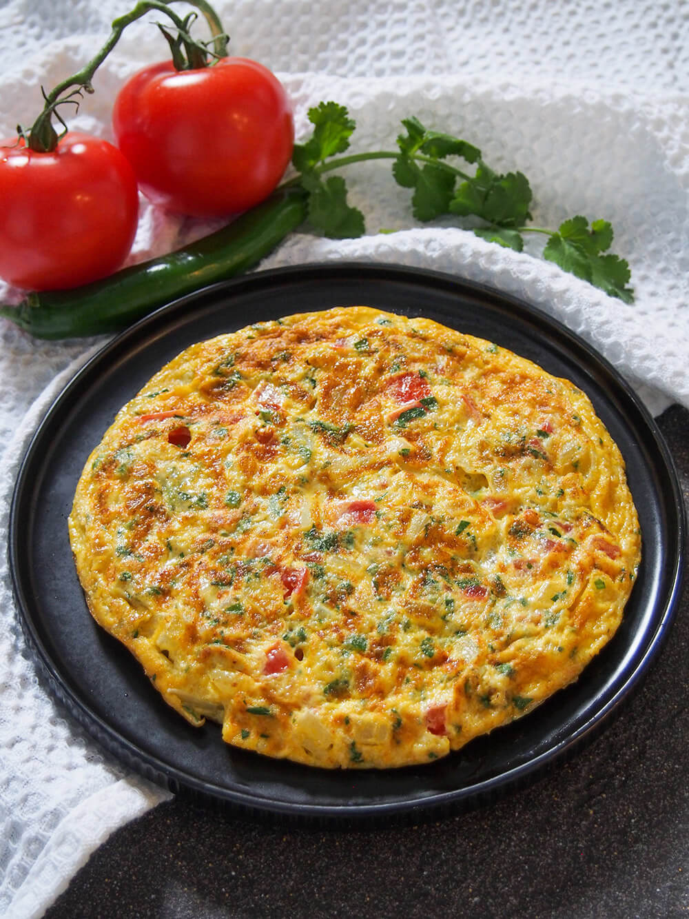 Masala omelette is a popular Indian breakfast packed with flavor. It's quick and easy to make, with a simple combination of vegetables and a bit of spice. Enjoy it as it is or wrapped in a roti. masala omelette on plate with tomato, chili and cilantro behind