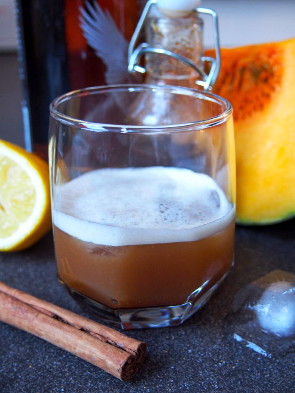 This pumpkin spice whiskey sour is the pumpkin spice cocktail you've been searching for! It's easy to make, with a wonderful balance of warm spices and tart lemon. A wonderful warm twist on a classic. glass of pumpkin spice whiskey sour with whiskey bottle, syrup bottle and pumpkin chunk behind