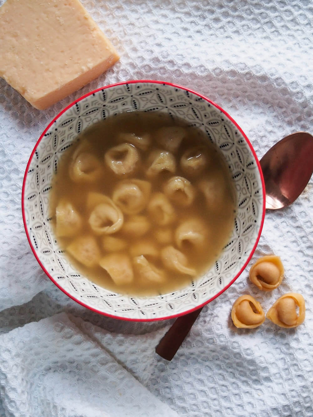 bowl of tortellini in brodo with spoon to one side and block of cheese above