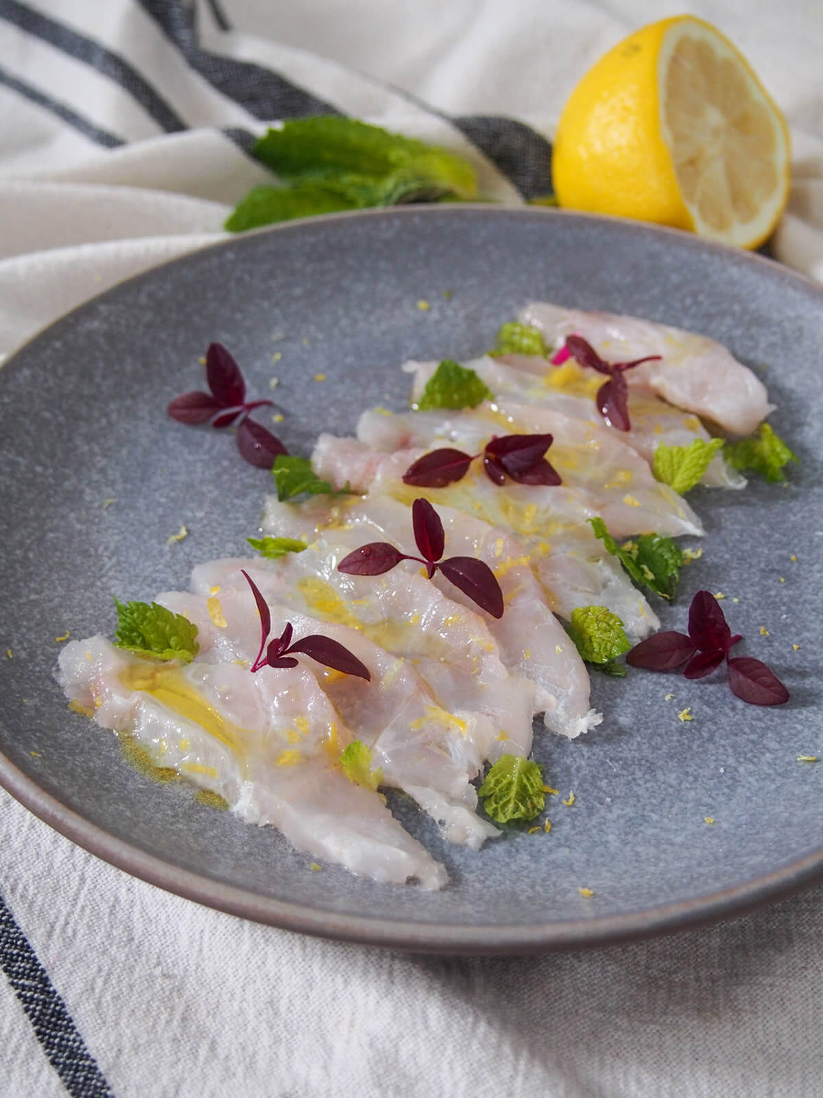 Fish crudo is a simple Italian preparation of raw fish. With only a few ingredients, it takes no time to prepare, but the result is elegant & delicious. plate of fish crudo with lemon and mint behind plate