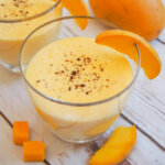 glass of mango lassi with mango garnish and mango behind