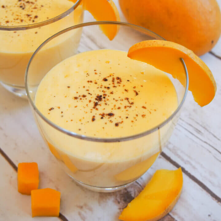 glass of mango lassi with mango garnish and mango behind