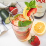 glass of pimm's cup cocktail with strawberries, cucumber and bottle around glass