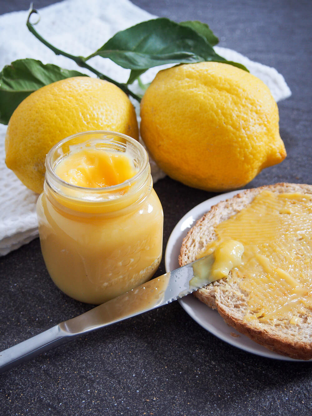 jar of lemon curd, with toast with some on to side and lemons behind