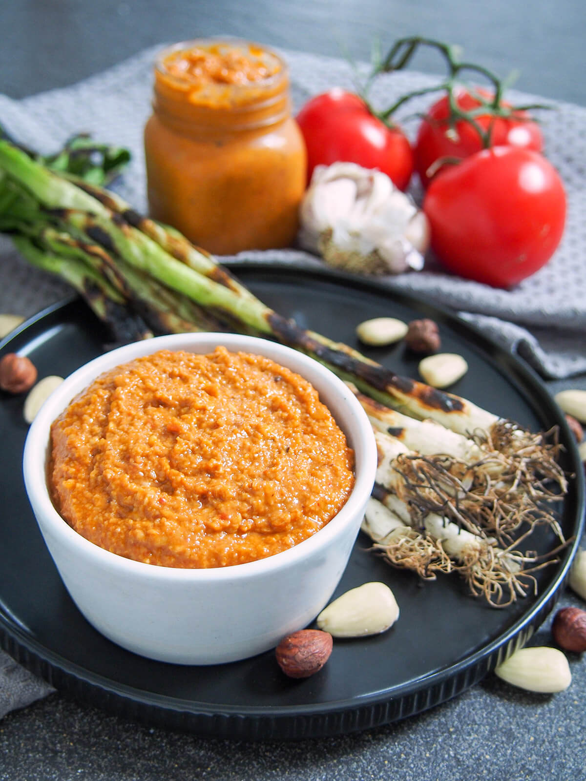 romesco sauce in dish with grilled spring onions behind and jar of sauce at back