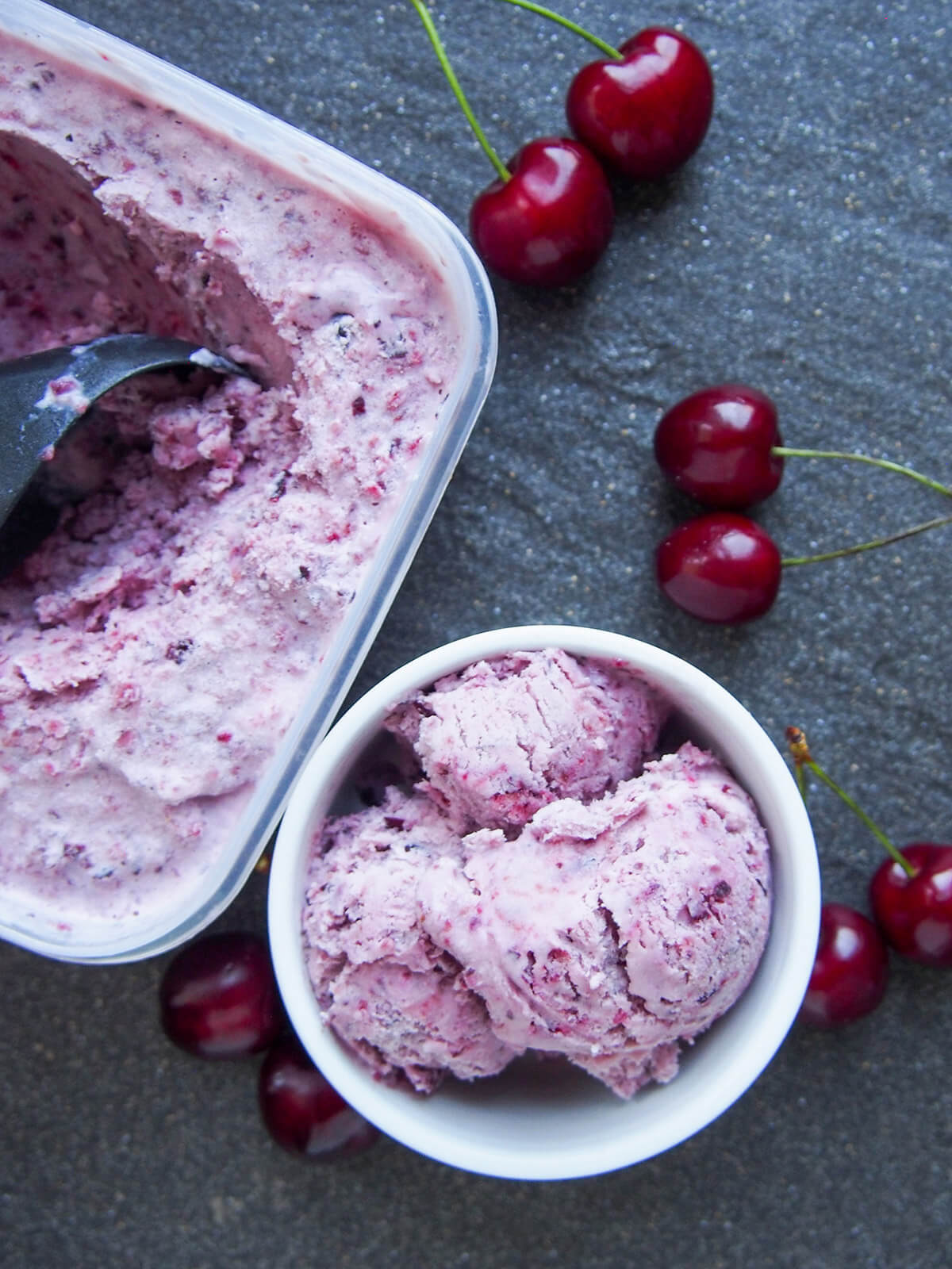 This cherry ice cream is easy to make and packed with wonderfully bright cherry flavor. It uses a simple no churn method that results in a wonderful balance of sweetness, creaminess and fruitiness. bowl of cherry ice cream with tub to side and cherries around both from overhead