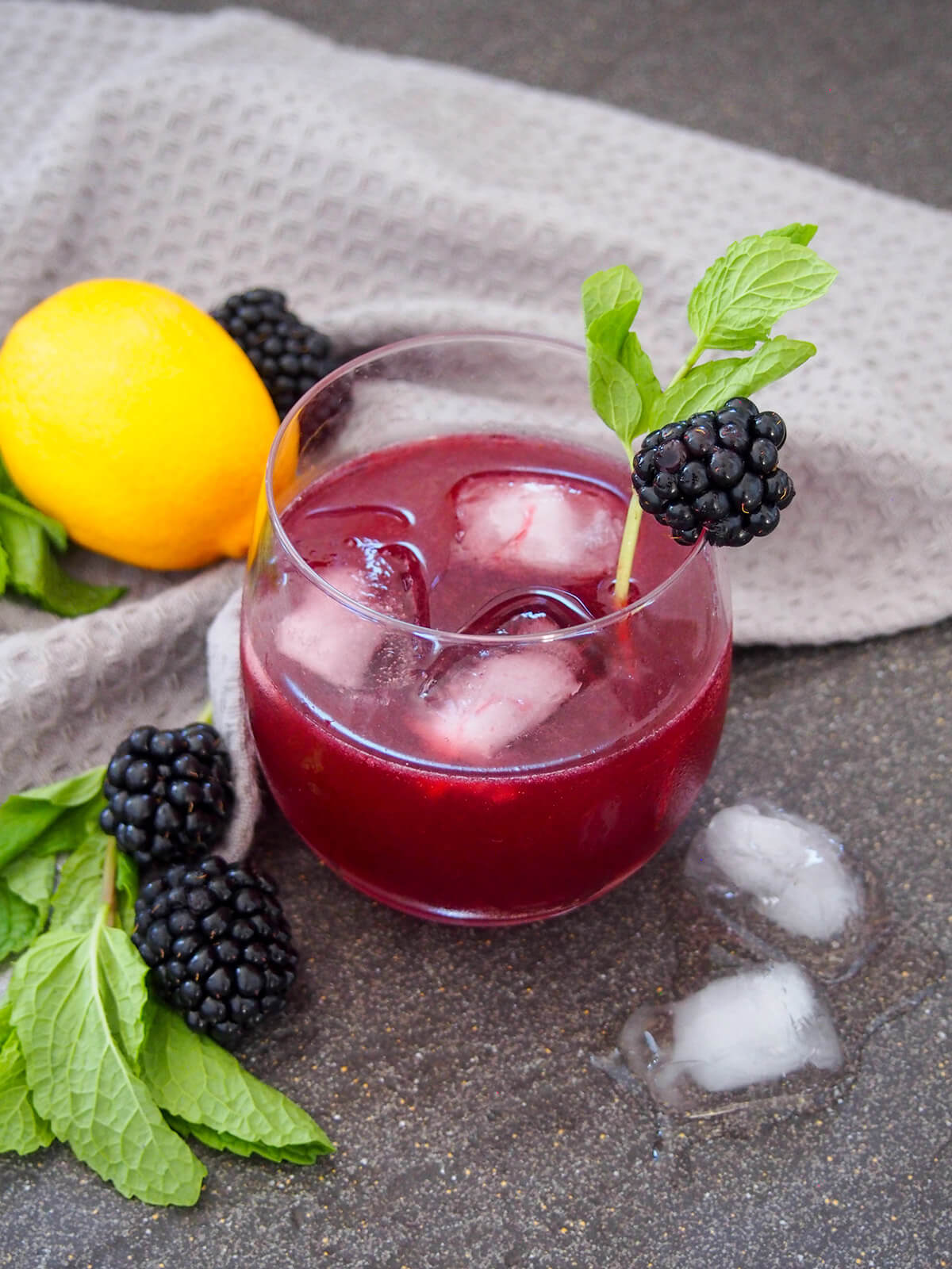 glass of blackberry bourbon smash with grey cloth behind and blackberries, lemon and mint to side