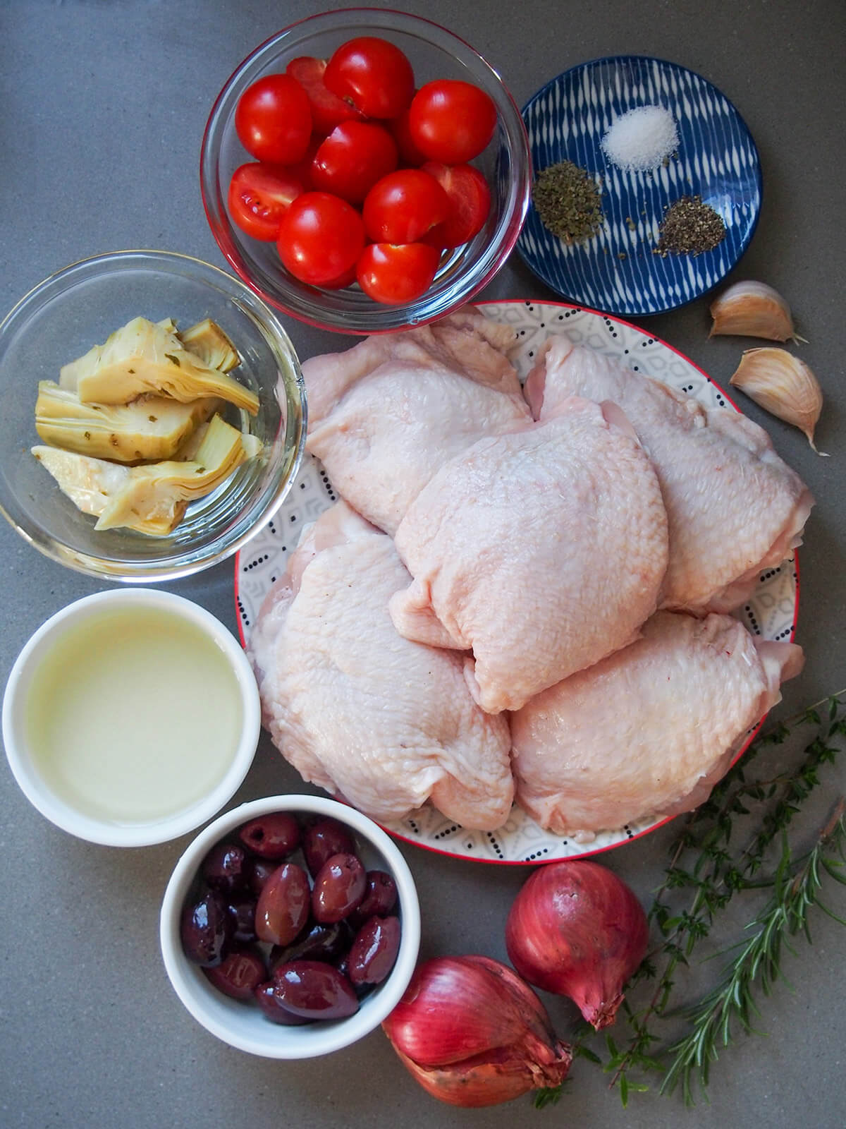 plate of chicken thighs with bowls with tomatoes, artichokes, wine, olives and herbs around it