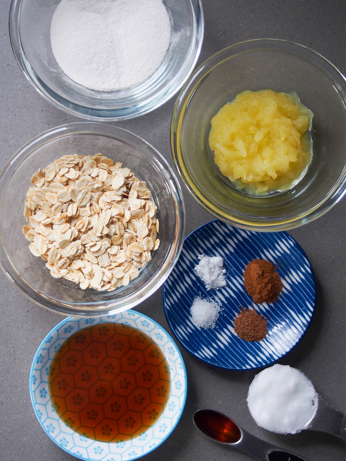 ingredients for spiced maple oatmeal cookies bowls with oats, applesauce, rice flour, maple, spices and spoons of vanilla and coconut oil