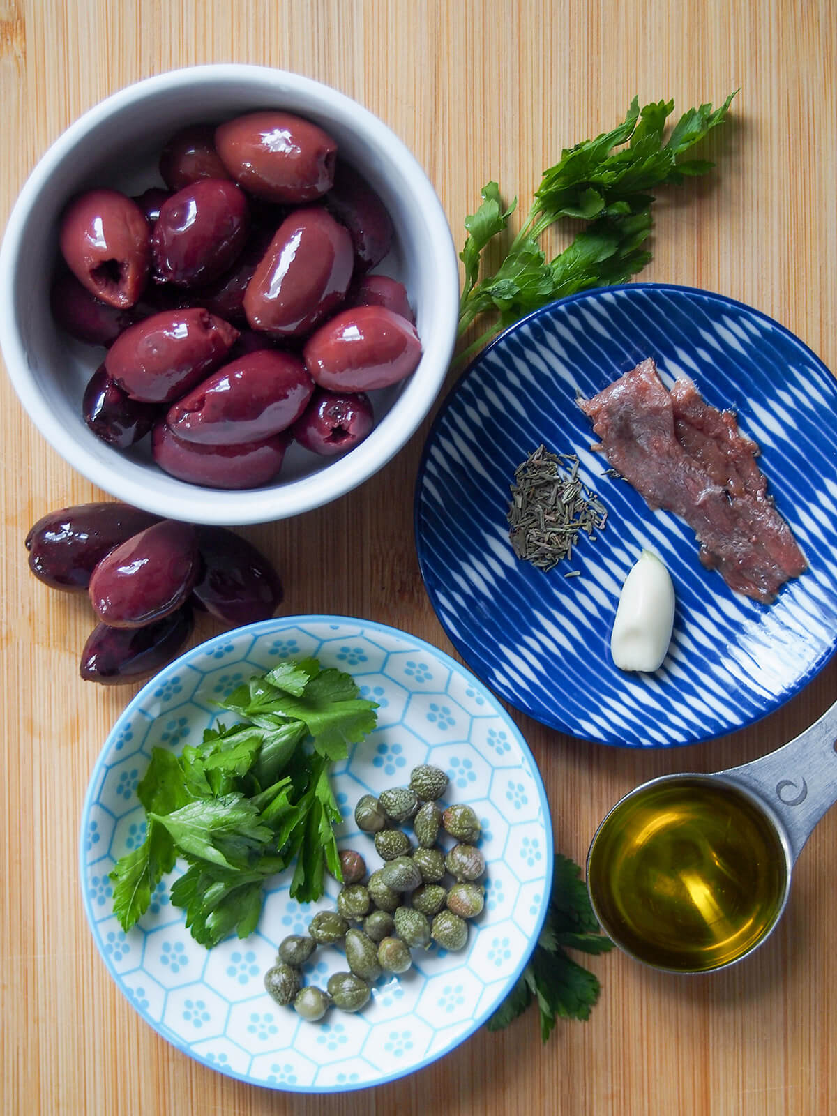 bowls with olives, capes, parsley, anchovies, garlic and thyme and spoon of olive oil to side