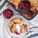 serving of plum crumble on plate with cream and rest of crumble in dish above