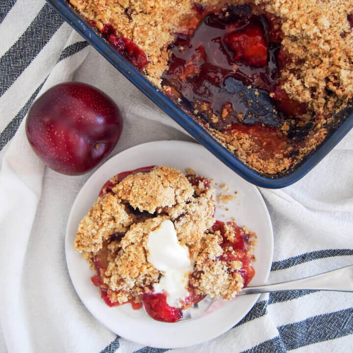 serving of plum crumble on plate with cream and rest of crumble in dish above